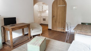 Una stanza con un tavolo di legno, una poltrona bianca, un piccolo sgabello di legno, una sedia di plastica trasparente e una porta in legno.