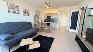 A modern living room with a grey sofa, a black rug, and a small wooden table. There is a kitchen area in the background with a dining table and chairs. - 06DO : Beau studio rénové face à la mer (Antibes)