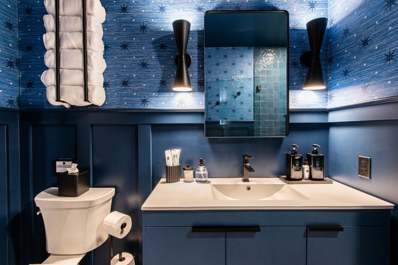 A bathroom with a large mirror, two wall-mounted lights, a toilet, and a sink with toiletries.