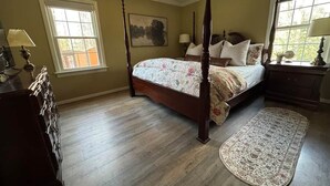 A bedroom with a four-poster bed, a wooden dresser, a nightstand, a window with a view, and a floor lamp.