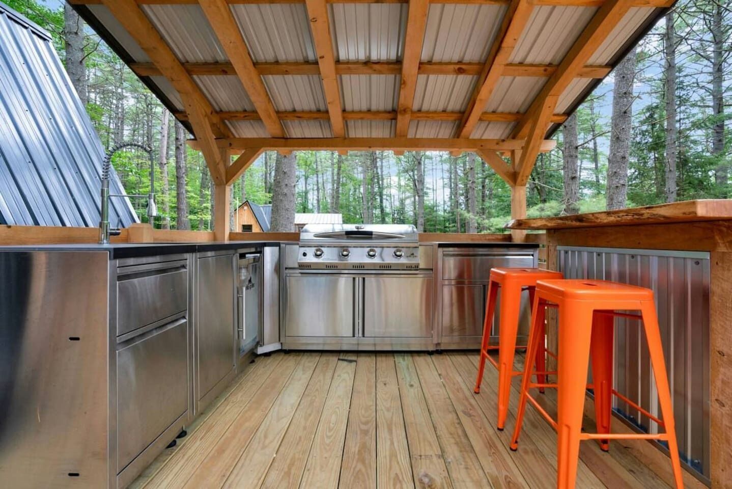 A covered outdoor kitchen with stainless steel appliances, wooden deck flooring, and a metal roof structure.