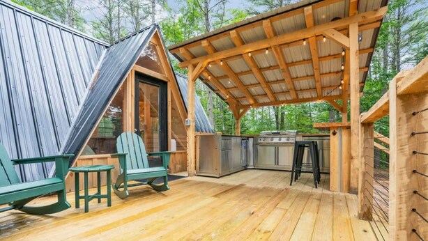 A wooden deck with a grill and bar stools, a kitchen area, and a covered seating area with a view of the surrounding forest.
