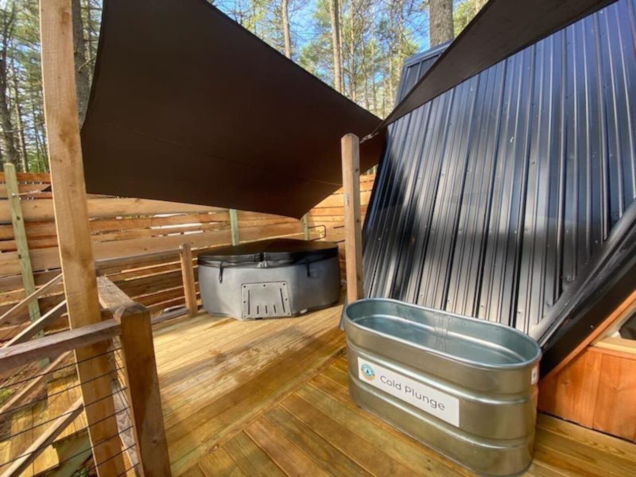 Une terrasse en bois avec un conteneur métallique portant l’inscription « Bain d’eau froide » et un jacuzzi noir.