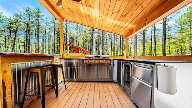 Terraza de madera con área de cocina, que incluye una parrilla, taburetes de bar y electrodomésticos de acero inoxidable, con vista a un bosque.