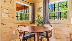 Interior de una cabaña de madera con una mesa redonda, dos sillas y ventanas amplias que dan a un bosque.