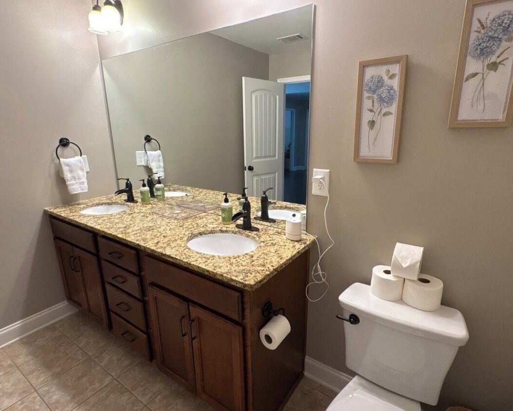 A bathroom with a granite countertop, two sinks, a toilet, a large mirror, and a framed floral artwork on the wall.