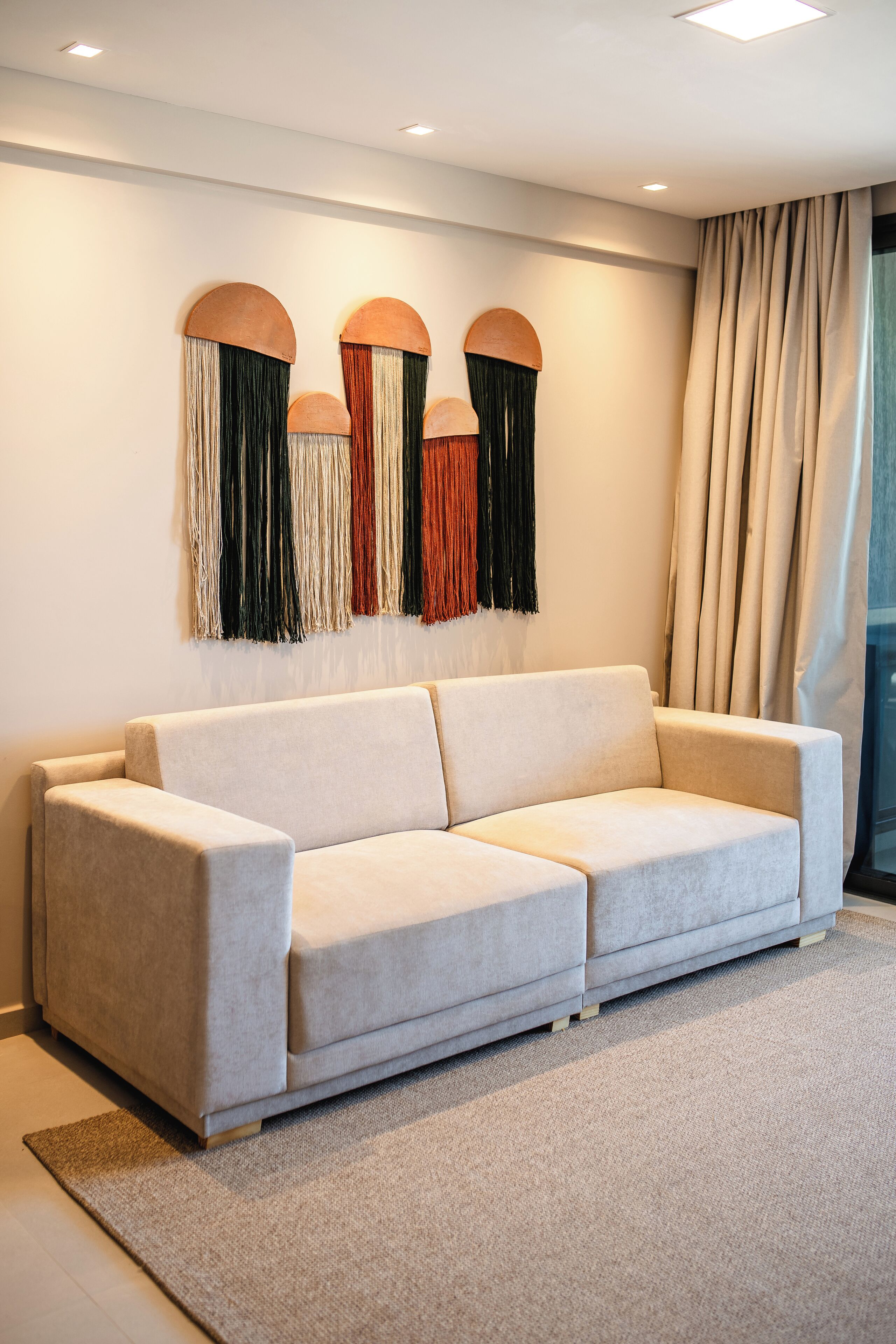 A modern living room with a light-colored sofa, a decorative wall hanging, and a large window with curtains.