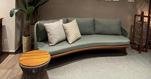 A curved green sofa with two pillows, a round wooden side table, and a woven floor lamp. - Praia Exclusive Muro Alto by Sintta (Ipojuca)