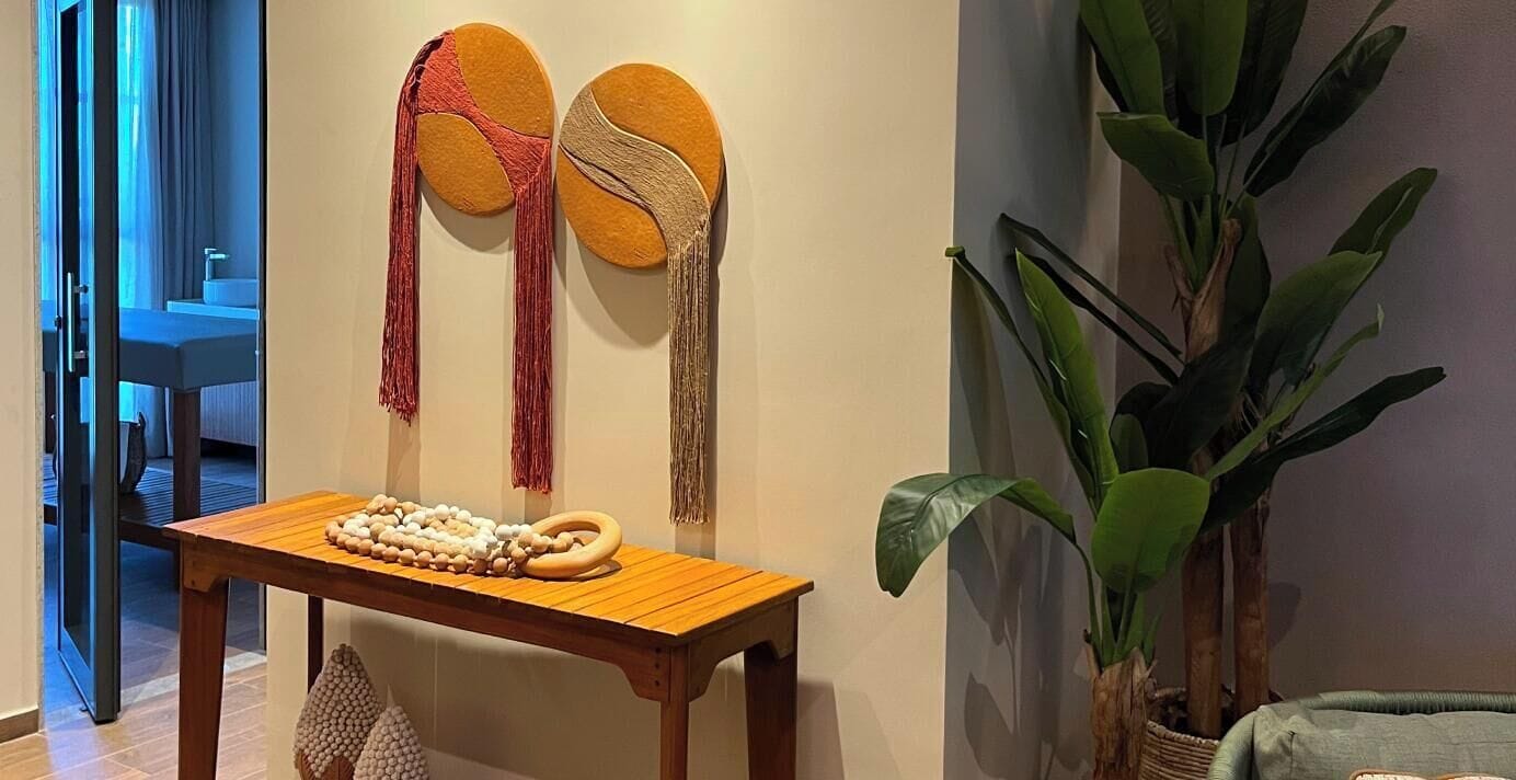 A wooden table with a string of wooden beads and a wooden ring, a large green plant, and two wall-mounted decorative items with tassels.