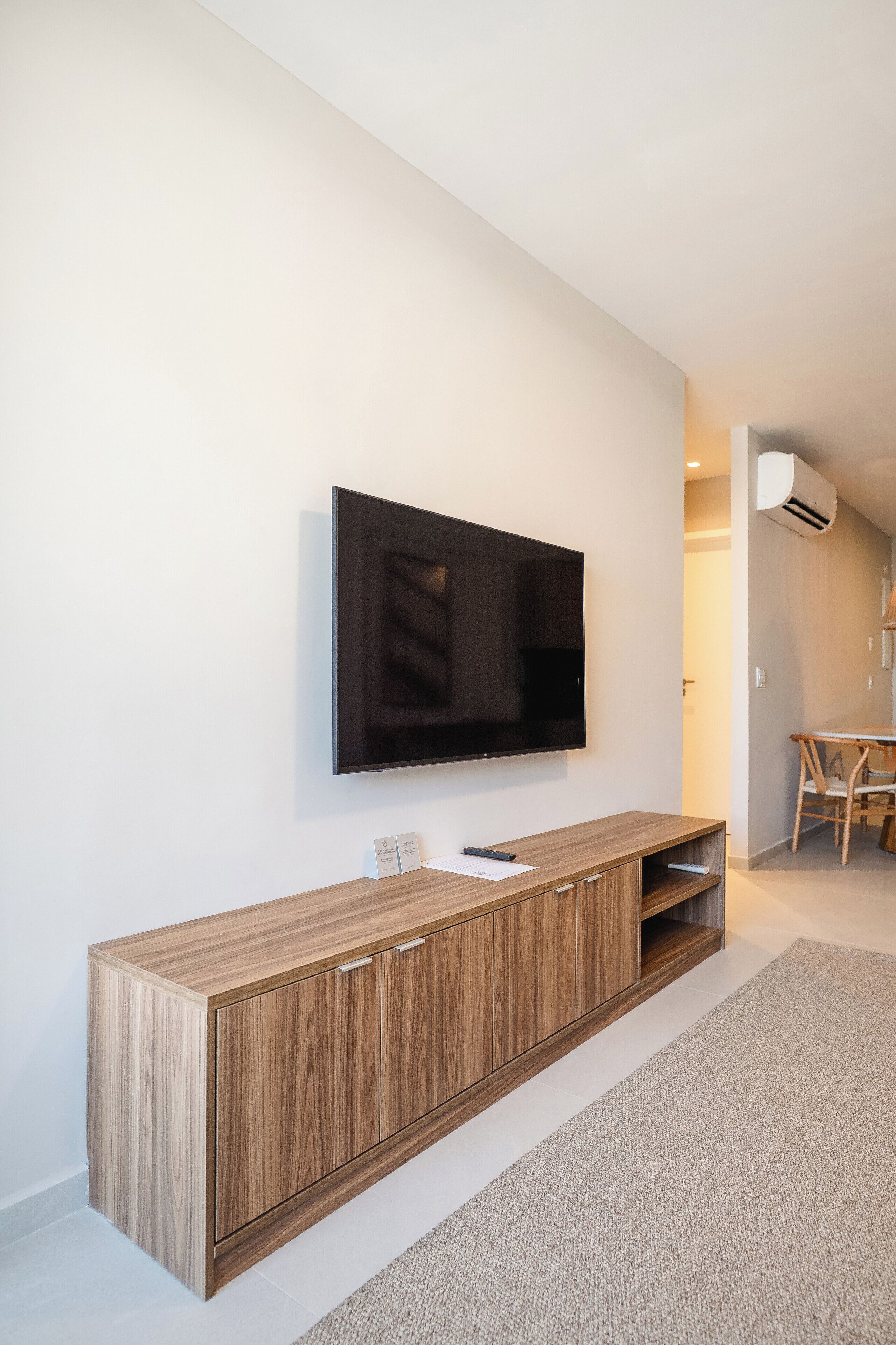 A modern living room with a flat-screen TV mounted on a wooden cabinet, a small table with chairs, and an air conditioning unit on the wall.