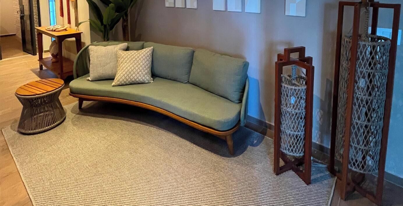 A curved green sofa with patterned cushions, a round wooden side table, and a woven floor rug in a room with a wooden floor.