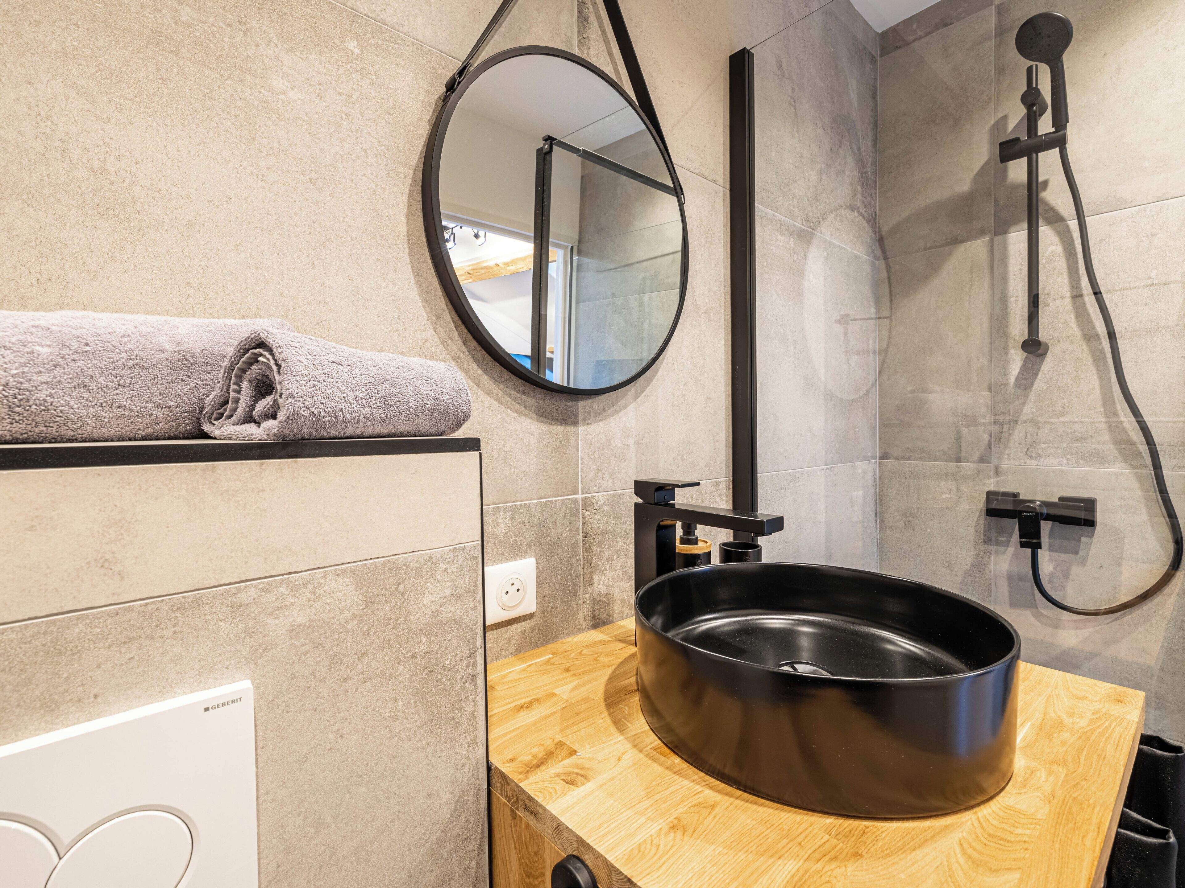 Un baño moderno con tocador de madera, un lavamanos negro, una toalla sobre la encimera, un espejo y un cabezal de ducha.