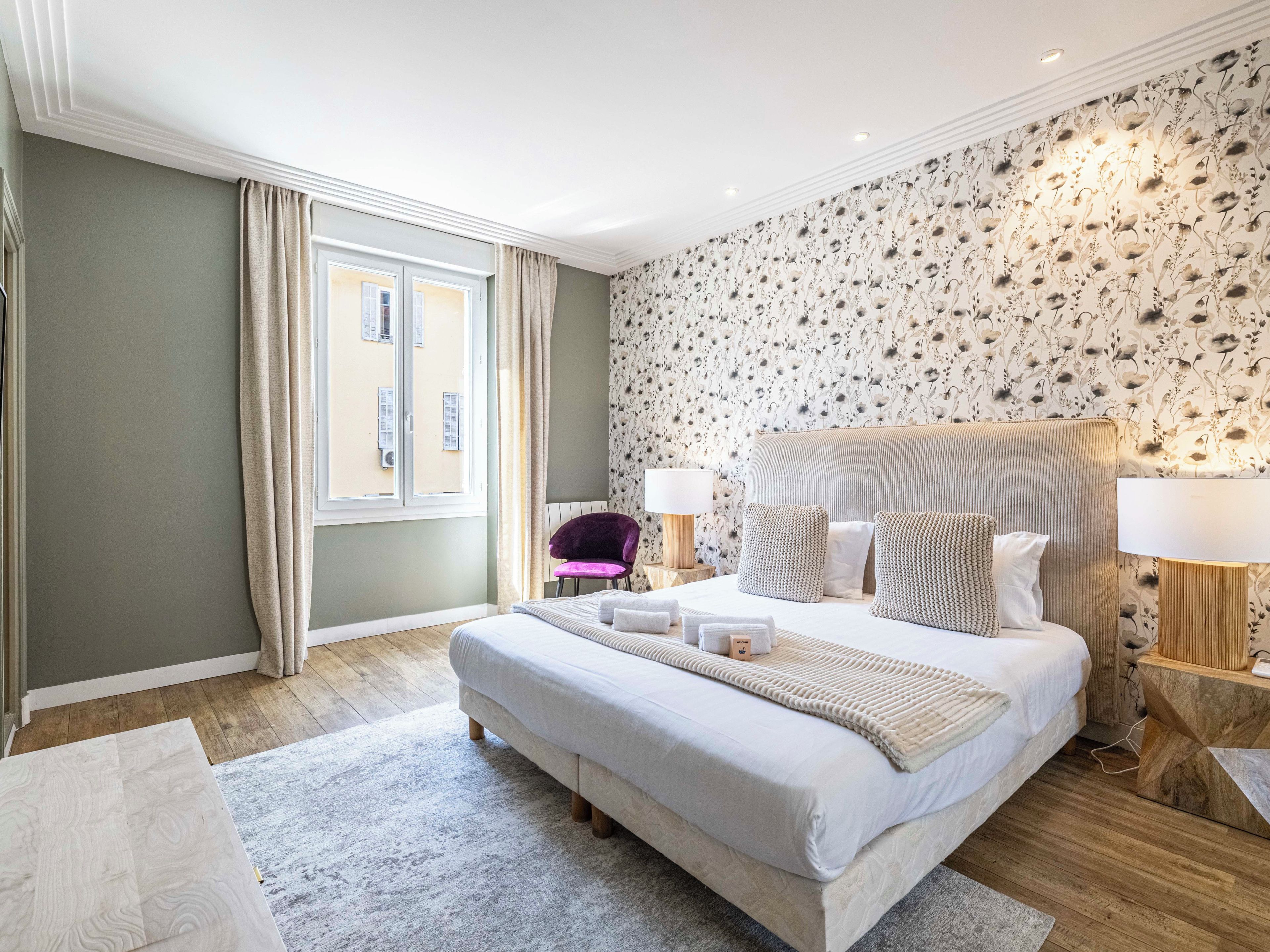 Ein Hotelzimmer mit einem großen Bett, zwei Nachttischlampen, einem Fenster mit Vorhängen, einer gemusterten Tapete und einem Holzboden.