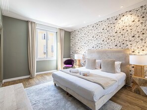 A hotel room with a large bed, two bedside lamps, a window with curtains, a patterned wallpaper, and a wooden floor. - 3-bedroom apartment Rue d'Antibes near the Palais (Cannes)