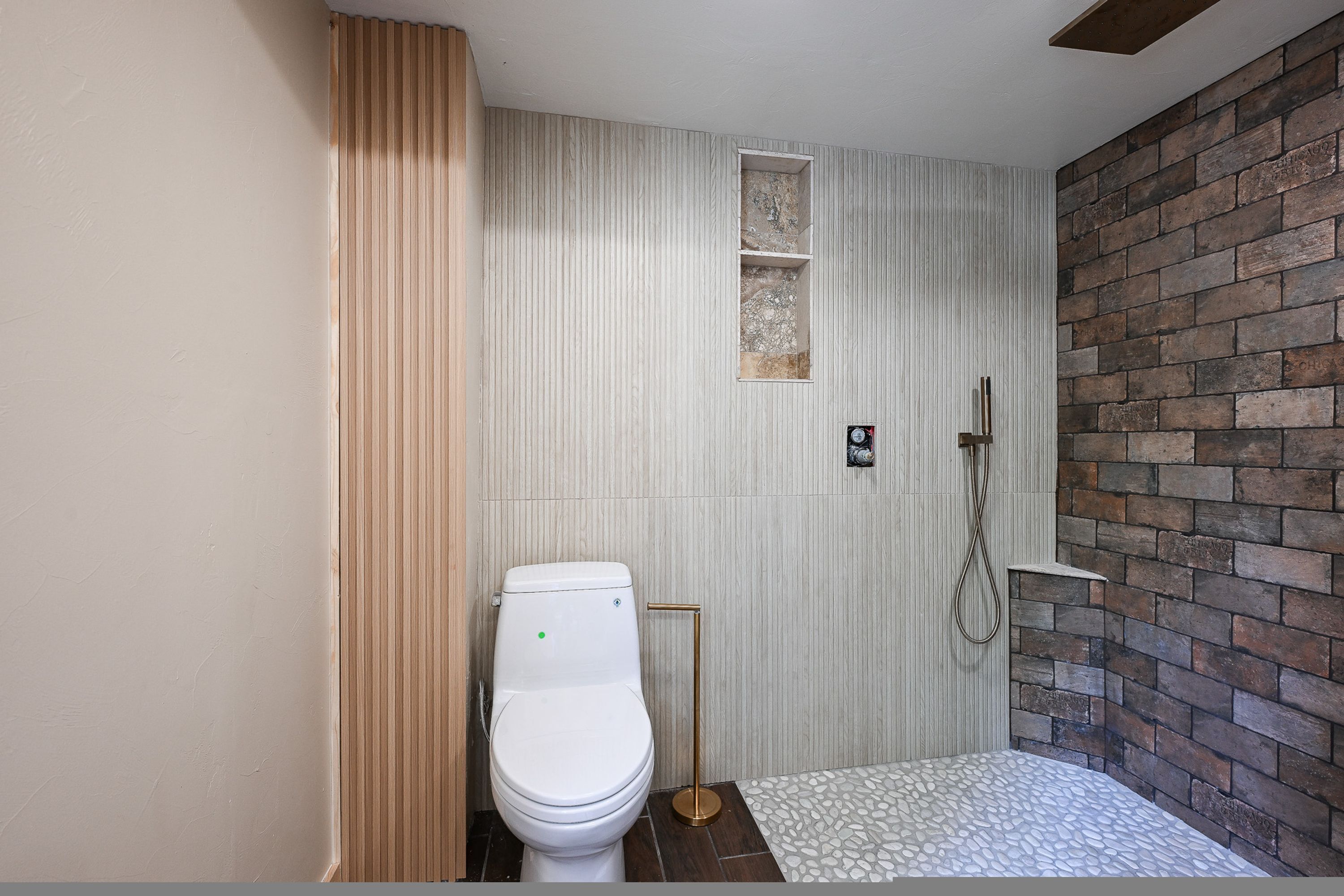 A bathroom with a white toilet, a shower with a wooden floor, and a brick wall.