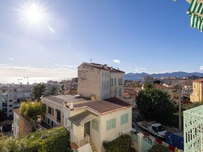 Un paysage urbain côtier avec un bâtiment imposant doté d’un balcon, offrant une vue sur la mer et les montagnes en arrière-plan.
