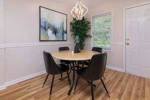 A dining area with a round table, black chairs, a chandelier, a painting, and a window with blinds. - Longer Stay 1BR with Comfortable Living & Workspace (Charlotte)