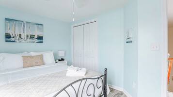 A bedroom with a bed, white bedding, a ceiling fan, a white door, and a painting of sailboats on the wall.