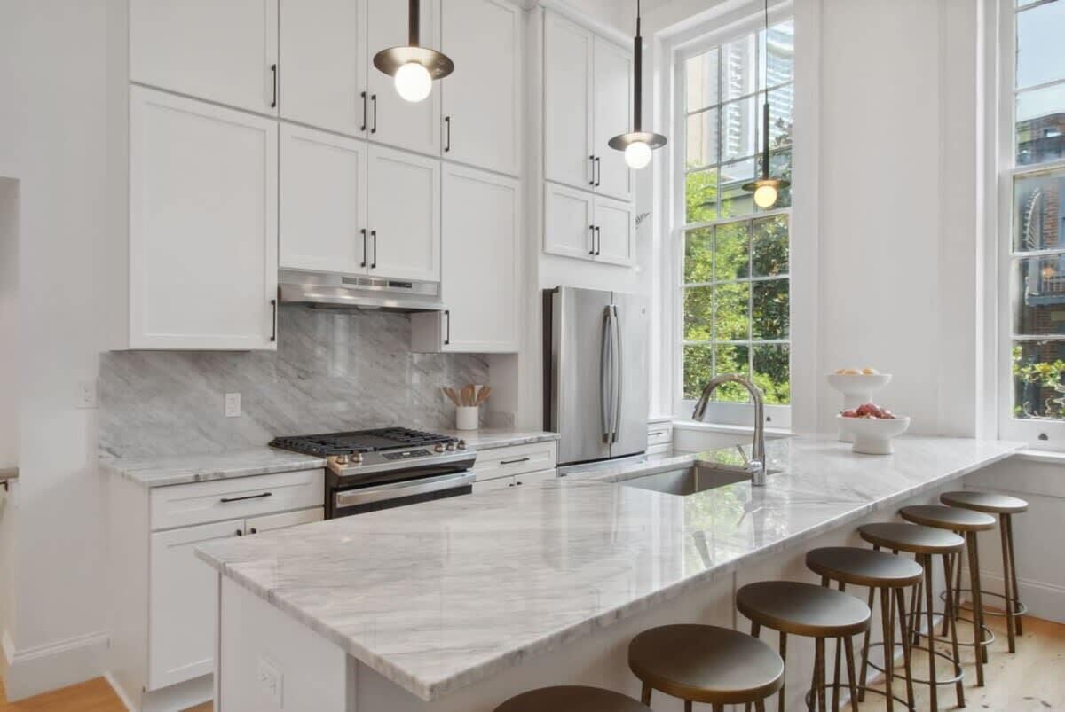 Una cocina moderna con armarios blancos, encimera de mármol, electrodomésticos de acero inoxidable y un amplio ventanal con vistas a la vegetación.