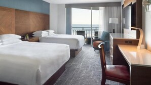 A hotel room with two beds, a desk, a chair, a TV, and a balcony with a view.