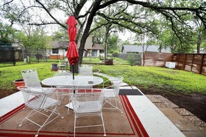 Un patio trasero con una mesa redonda y sillas, una sombrilla roja y un árbol.