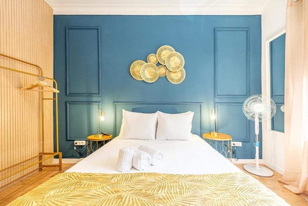 Una camera d'albergo con un letto grande, due comodini, un oggetto decorativo a parete e una ventola da terra.