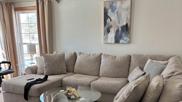 Ein Wohnzimmer mit einem beigefarbenen Ecksofa, einem Glastisch, einem gemusterten Teppich und einem modernen abstrakten Gemälde an der Wand.