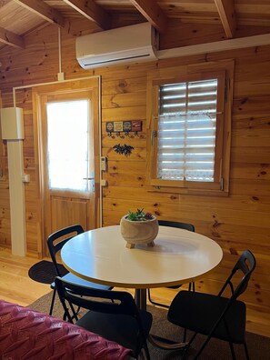 L’intérieur d’un chalet accueillant, avec une table ronde, deux chaises, une plante en pot, des murs en bois, une porte et une fenêtre avec des stores.