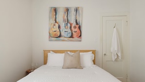 A bedroom with a bed, pillows, a painting of guitars, a door with a towel, and a nightstand with glasses. - Historic Uptown New Orleans 2BR – 1 Block to St. Charles Streetcar (New Orleans)
