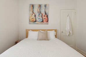 A bedroom with a bed, pillows, a painting of guitars, a door with a towel, and a nightstand with glasses. - Historic Uptown New Orleans 2BR – 1 Block to St. Charles Streetcar (New Orleans)