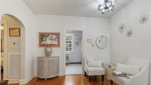 A spacious room with hardwood flooring, a white sofa, a white armchair, a side table with a magazine, a decorative vase, and a framed picture on the wall. - Historic Uptown New Orleans 2BR – 1 Block to St. Charles Streetcar (New Orleans)