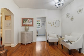A spacious room with hardwood flooring, a white sofa, a white armchair, a side table with a magazine, a decorative vase, and a framed picture on the wall. - Historic Uptown New Orleans 2BR – 1 Block to St. Charles Streetcar (New Orleans)