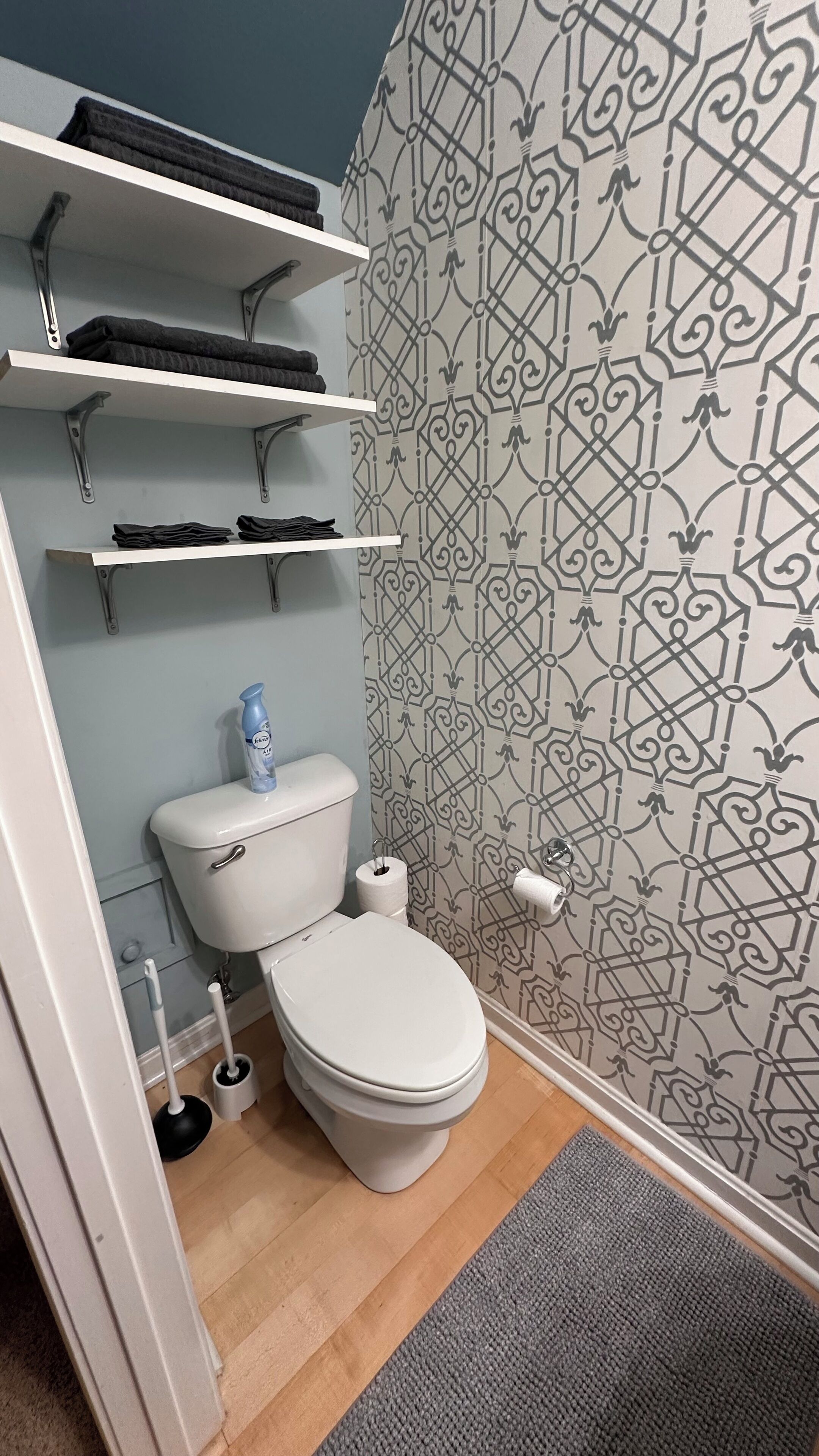 A bathroom with a toilet, a shelf with folded towels, a toilet paper holder, and a patterned wall.