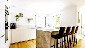 A modern kitchen with white cabinets, a large island with bar stools, and a window providing natural light. - Villa, close to beaches (Pornic)