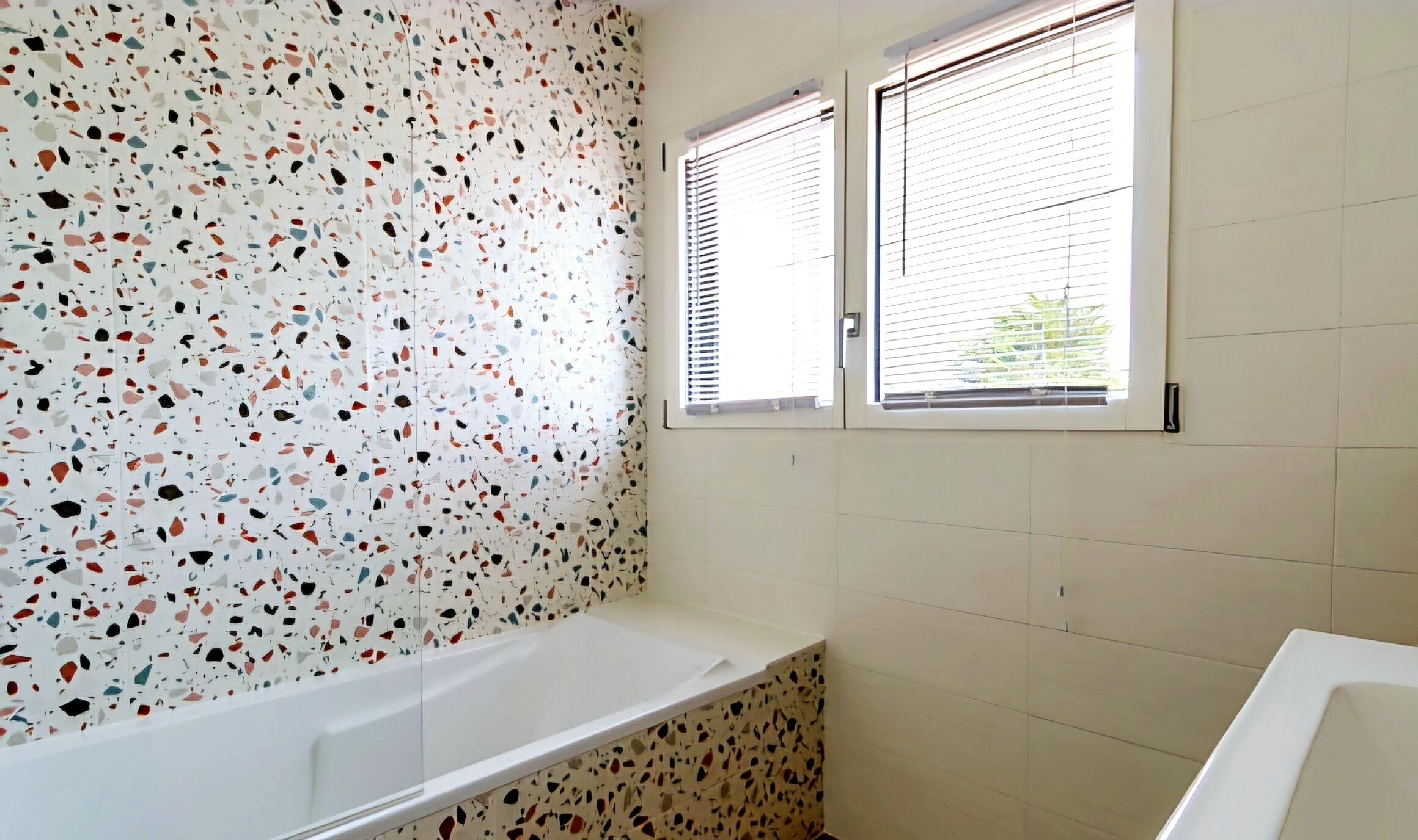A bathroom with a white bathtub, terrazzo flooring, and two windows with blinds.