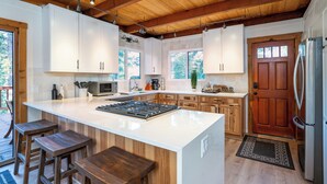 A modern kitchen with white cabinets, a large island with stools, a stainless steel refrigerator, and wooden beams on the ceiling. - Lakeview, Beautifully Remodeled Hilltop Cabin With Wraparound Deck. (Carnelian Bay)