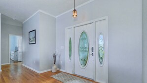 Un pasillo con suelo de madera, una puerta blanca con paneles de vidrio verde, un cuadro enmarcado en la pared y una lámpara colgante.