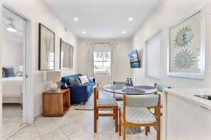 Ein modernes Wohnzimmer mit einem blauen Sofa, einem runden Esstisch mit Stühlen, einem kleinen Holznachttisch und einem an der Wand montierten Fernseher.