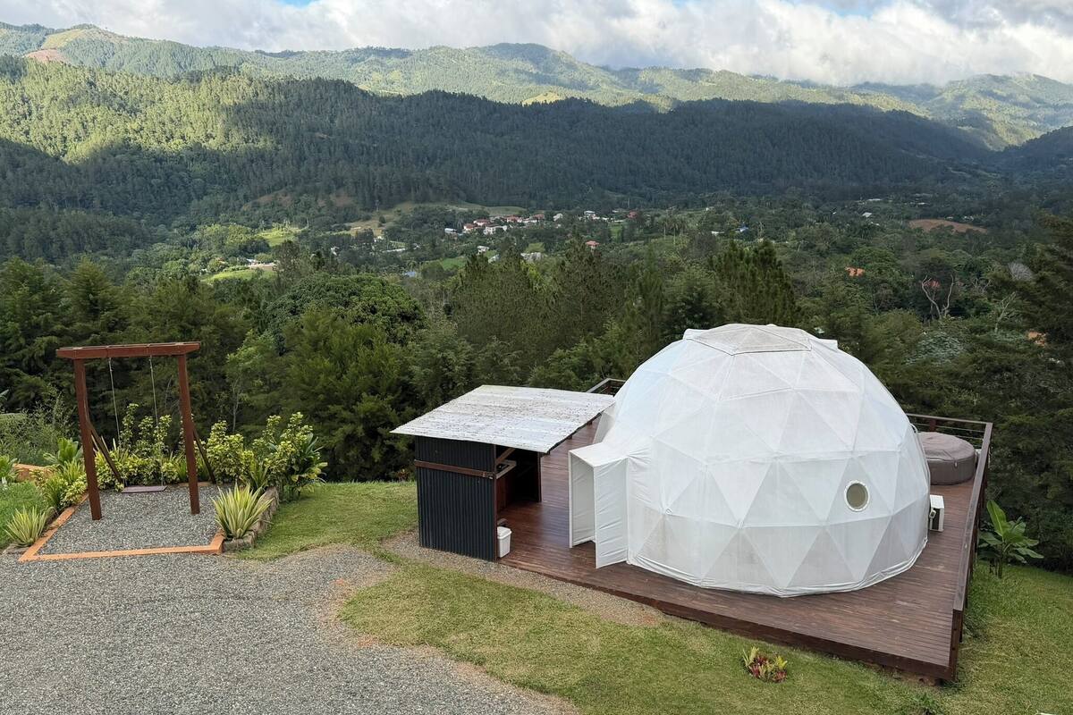 Una estructura de domo geodésico con un muelle de madera, rodeado por un jardín con un pérgola, con montañas al fondo.