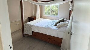 Un dormitorio con estructura de cama de madera, ropa de cama blanca y ventana con vista a follaje.