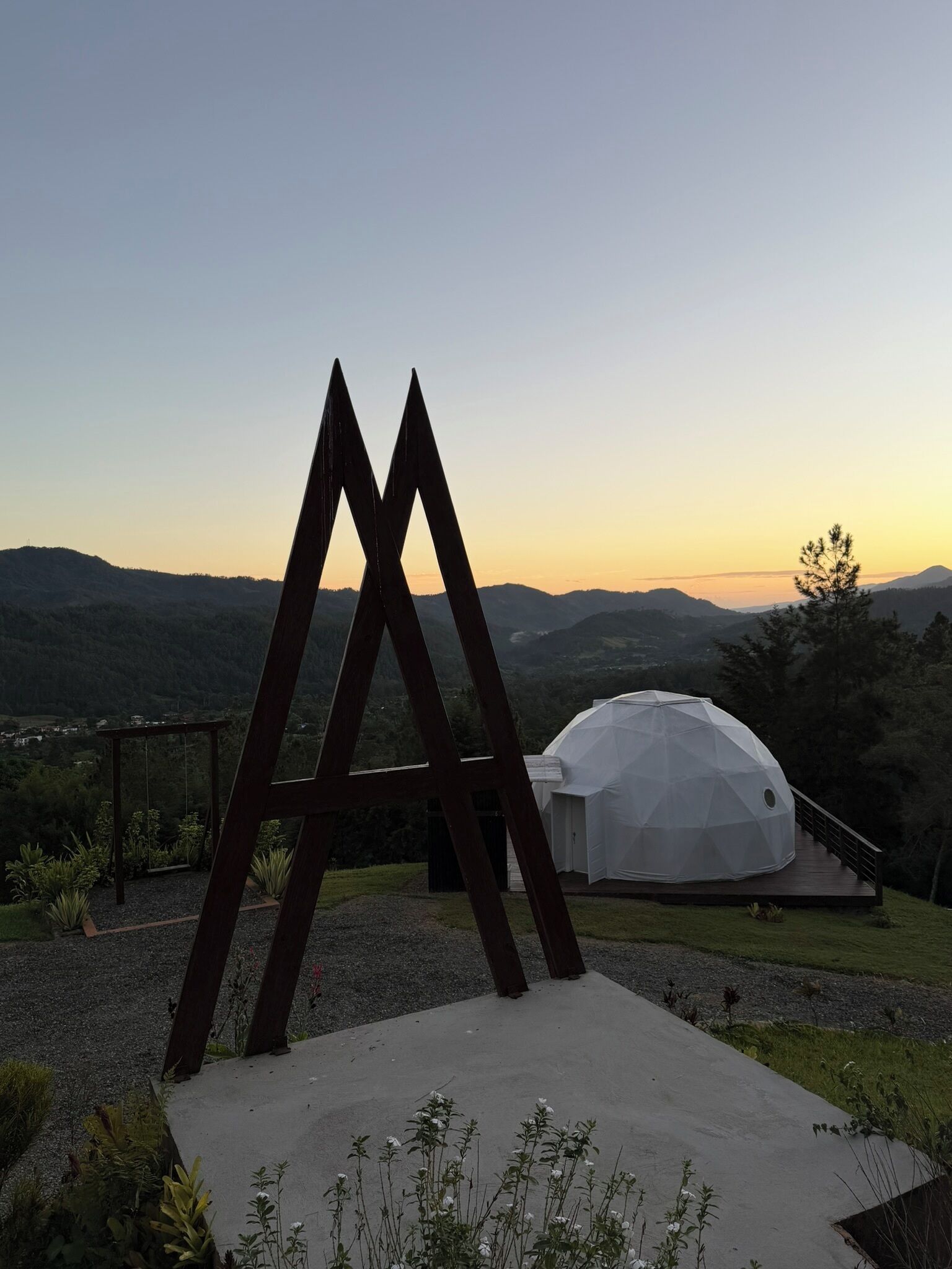 Una moderna estructura en forma de cúpula geométrica con una terraza de madera y una escultura metálica en primer plano, frente a colinas ondulantes y un cielo al atardecer.
