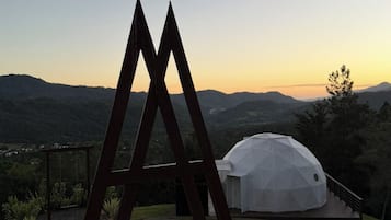 Une structure moderne en forme de dôme géodésique, dotée d'une terrasse en bois et d'une sculpture métallique en premier plan, le tout se détachant sur un paysage de collines ondulantes et un ciel au coucher du soleil.