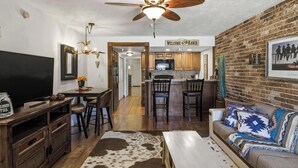 A living room with a TV, a sofa, a coffee table, and a fireplace. There is a kitchen in the background with a dining area. - River Ranch  Oasis /  Wild West Escape (River Ranch)