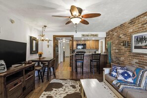 A living room with a TV, a sofa, a coffee table, and a fireplace. There is a kitchen in the background with a dining area. - River Ranch  Oasis /  Wild West Escape (River Ranch)