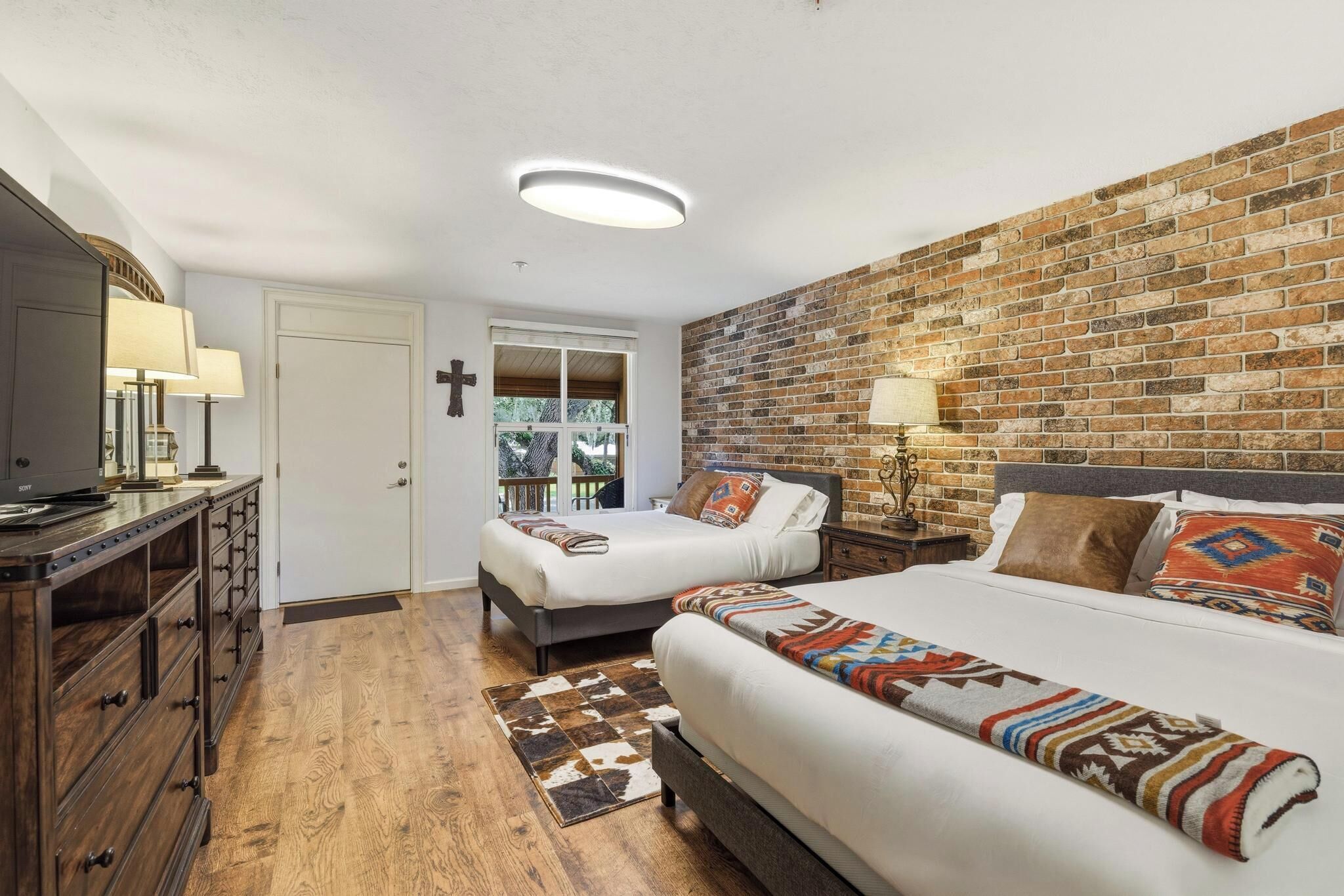 A bedroom with two beds, a dresser, a mirror, a cross on the wall, and a brick accent wall.
