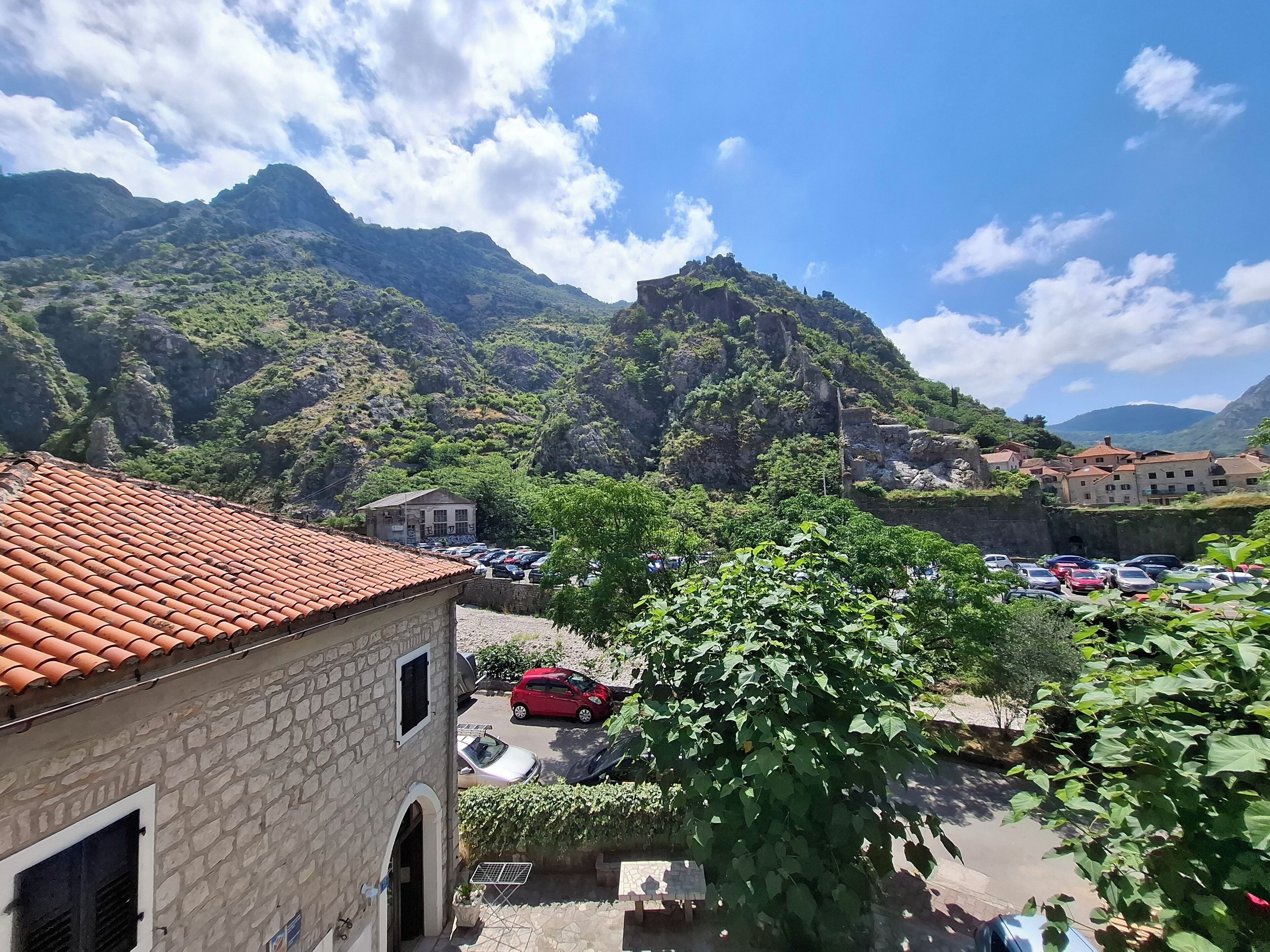 Un edificio de piedra con techo de tejas, un estacionamiento y montañas al fondo.
