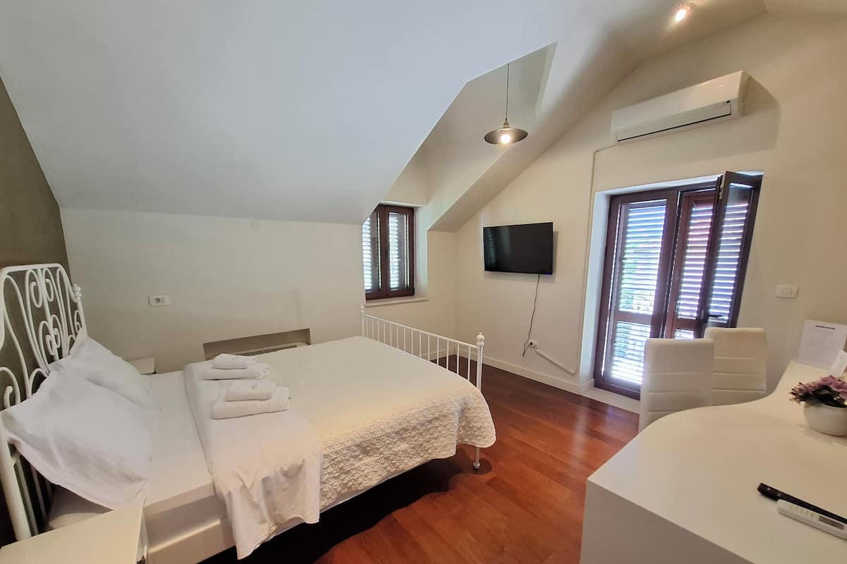 A bedroom with a bed, a desk, a chair, a TV, and a window with blinds.