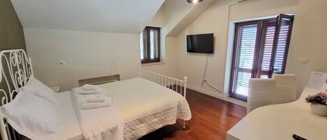 A bedroom with a bed, a desk, a chair, a TV, and a window with blinds.