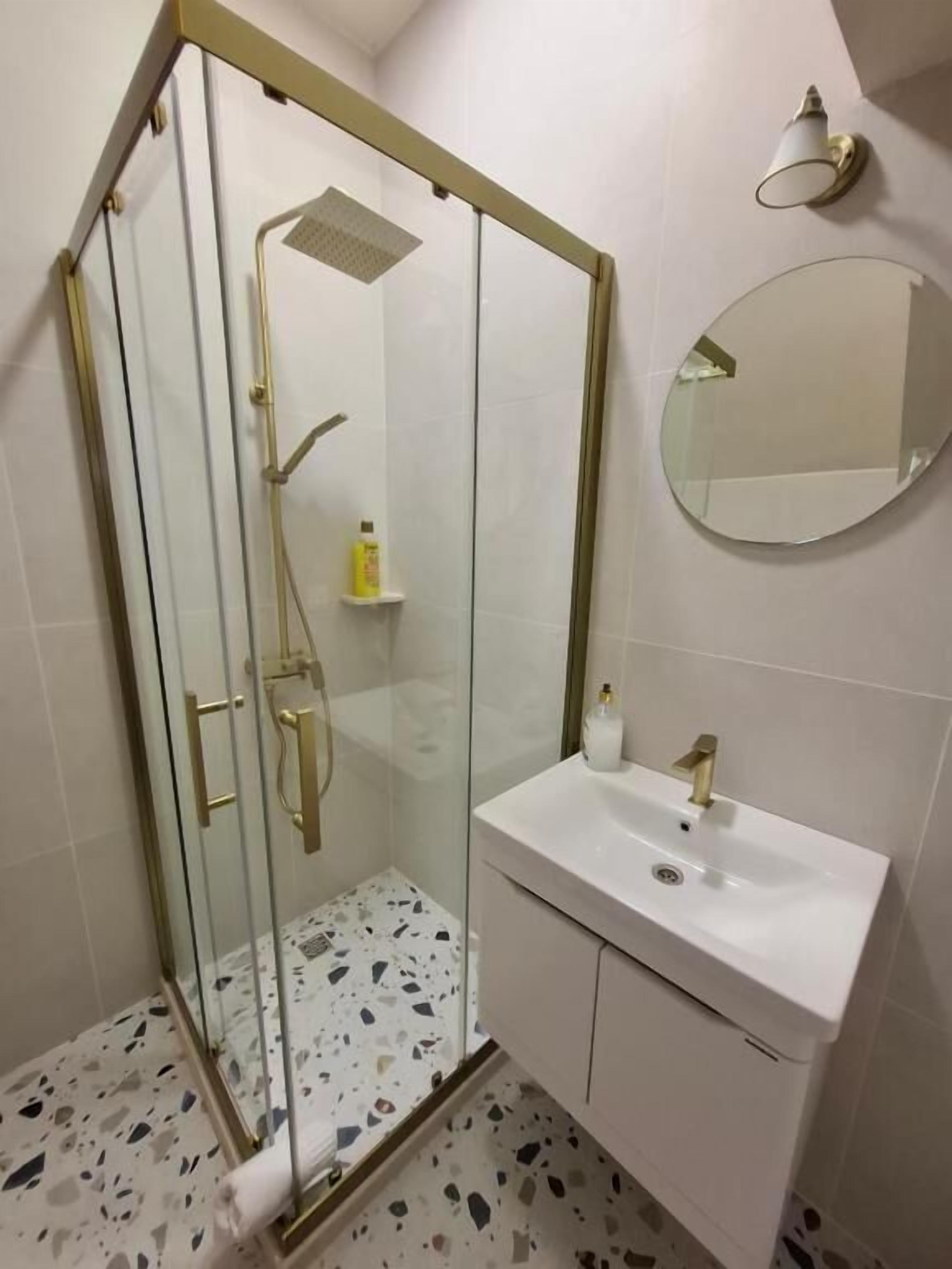 A modern bathroom with a glass-enclosed shower, a white sink with a gold faucet, and a round mirror on the wall.