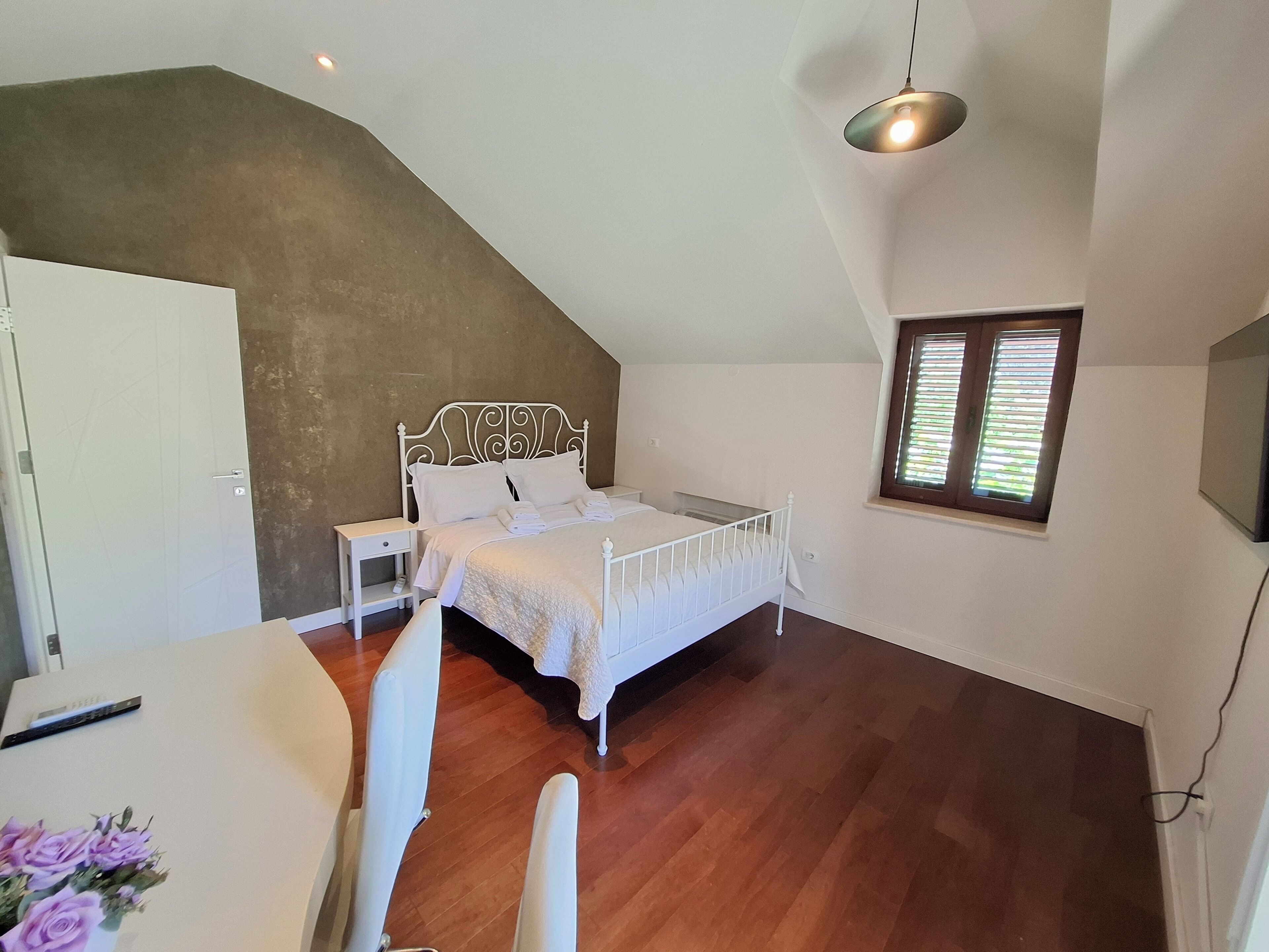 A bedroom with a sloped ceiling, a bed, a bedside table, a nightstand, a window with blinds, and a hanging light fixture.